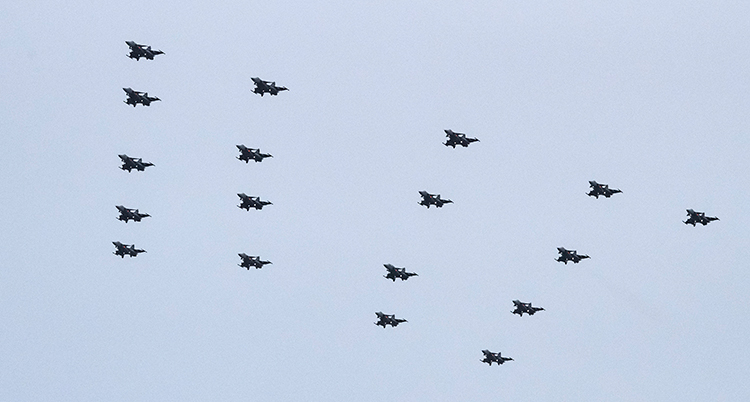 18 flygplan flyger i en formation likt en gran högt uppe i luften. Himlen är grå.
