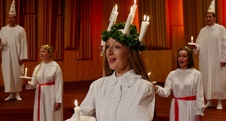 En lucia med en krona med ljus på huvudet, bakom syns tärnor i vita linnen och med ljud i händerna
