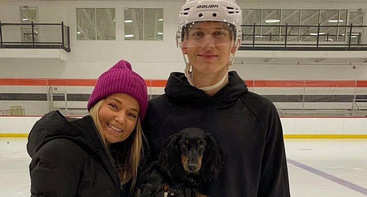 En kvinna med lila mössa lutar huvudet mot axeln på en man i svart jacka och vit hockeyhjälm. Båda tittar rakt in i kameran.