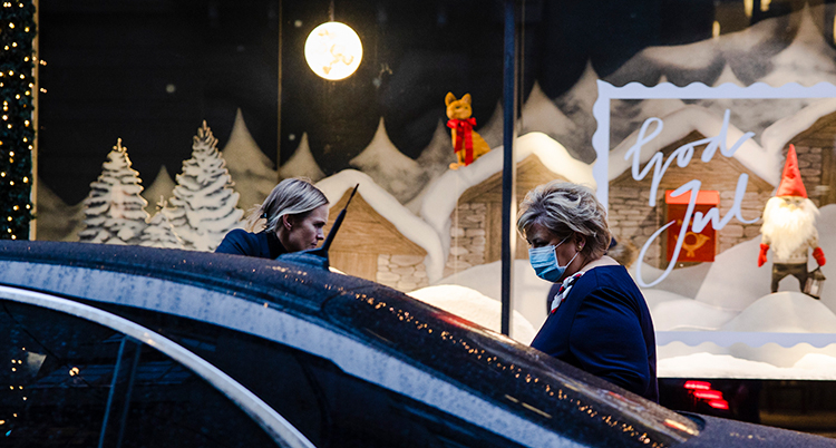 Hon står vid sidan av bilen. Hon har munksydd. I bakgrunden syns ett skyltfönster med en tomte i.