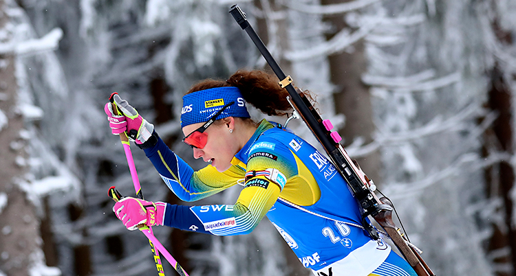 Hanna Öberg åker skidor. Hon har ett gevär på ryggen. Hon har en dräkt som är blå och gul. Och hon har glasögon med röda glas.