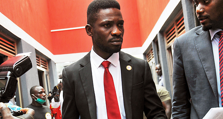 En bild på politikern Bobi Wine. Han har en mörk kostym, vit skjorta och en röd slips. Journalister vill prata med honom.