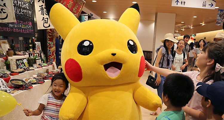 En person har klätt ut sig till figuren Pikachu. Pikachu är gul och har röda kinder. Flera barn vill leka med Pikachu. Bilden är tagen i Japan.