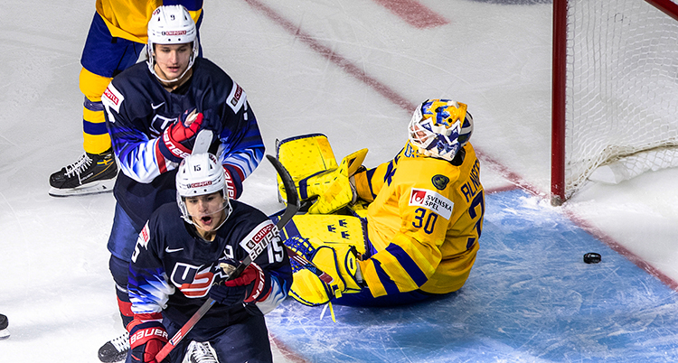 Sveriges målvakt sitter på isen. Pucken 'r bakom honom. Två amerikaner åker därifrån.