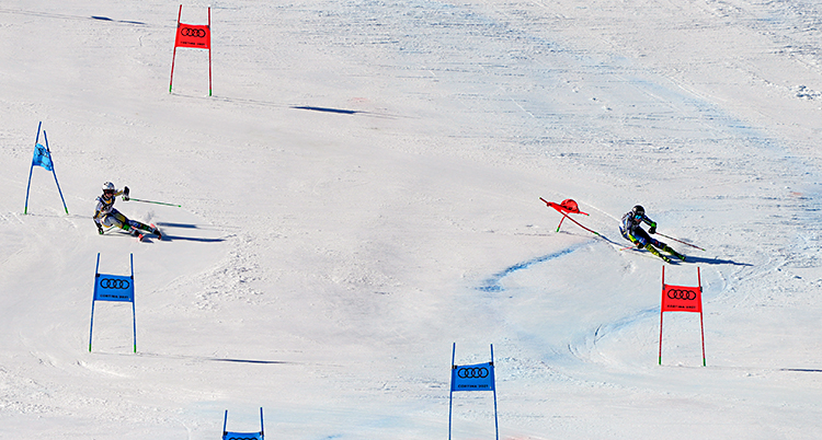 Två personer åker slalom bredvid varandra. Personen till vänster åker runt blå portar. Personen till höger åker runt röda portar.