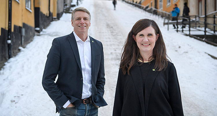De står utomhus på en gata. Det är snö på gatan. Per Bolund står till vänster. Han har kort ljust hår. Märta Stenevi står till höger. Hon har långt mörkt hår.