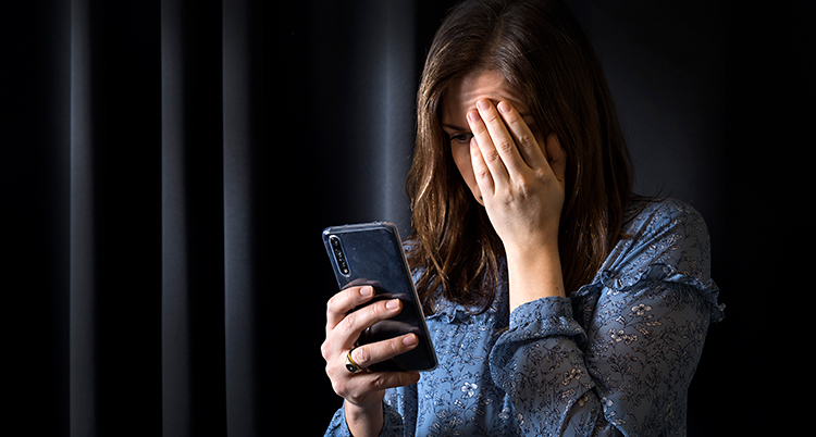 En kvinna står i ett rum. Hon håller i en mobiltelefon med ena handen. Den andra handen håller hon för ansiktet.