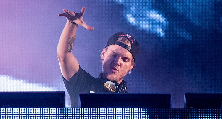 Avicii tittar neråt på en dator. Han står i blått och rosa ljus. Han har keps och hörlurar runt halsen. Han höjer ena handen och spretar med fingrarna.