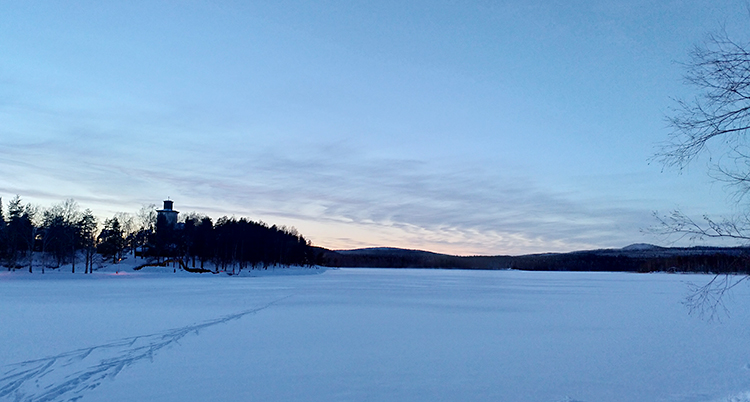 Solen håller på att gå ner över en älv. Det är snö på älven. Längre bort ser vi en skog.