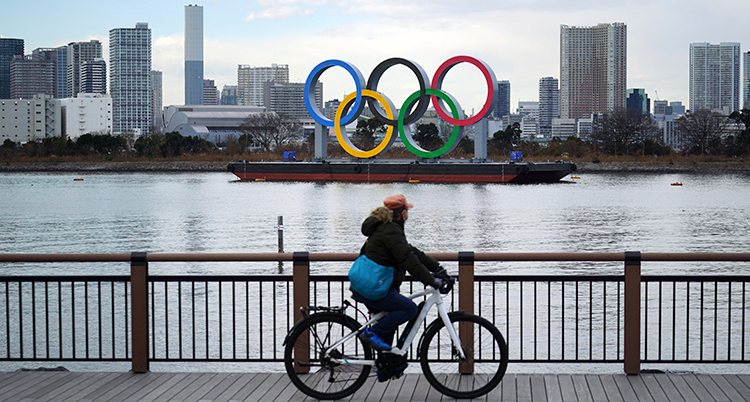 En man cyklar framför en flod. I bakgrunden syns de fem olympiska ringarna.