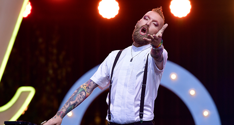 Han sträcker ut vänster arm framför sig, blundar och sjunger. Han har vit kortärmad skjorta och tatueringar som syns på armarna.
