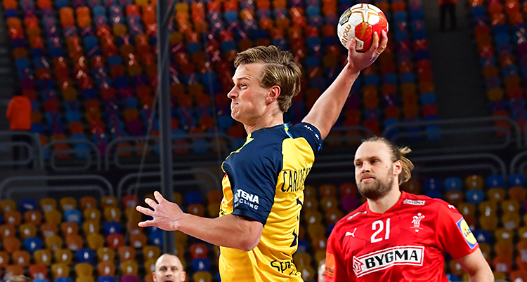 En match i handboll. En spelare i Sveriges lag håller i bollen. Han är på väg att skjuta. Han har gul tröja. Det andra laget har röda tröjor.
