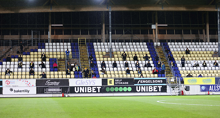 Bilder visar en läktare på en fotbollsarena. På läktaren är det få personer. De har flera tomma platser mellan sig.