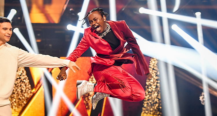 Bilden visar scenen i Melodifestivalen. Det är massor av strålkastare på scenen. Artisten Tusse har precis fått veta att han vunnit. Han hoppar upp i luften och skriker av glädje. Han har en röd kostym. Till vänster om Tusse står Eric Saade.