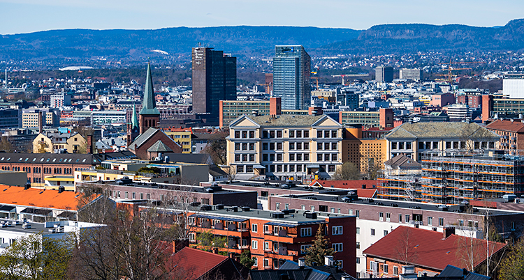 En bild av staden Oslo. Bilden är tagen från en hög byggnad. Vi ser en massa hus. I bakgrunden syns kullar. Solen skiner.