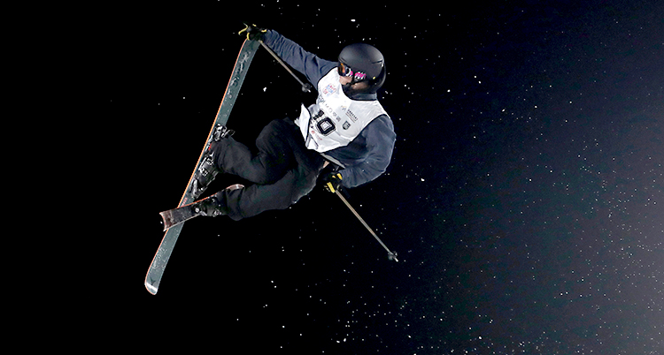 Oliwer Magnusson hoppar med skidor. Han är i luften. Han gör ett trick med skidorna. Han har svarta kläder och en svart hjälm.