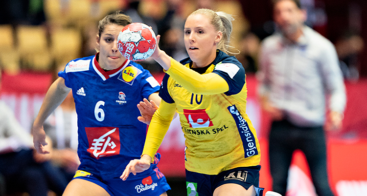 Från en match i handboll. En kvinna springer med bollen. Hon har ljust hår. Hon har gul tröja och blå shorts. En annan kvinna i blå kläder försöker ta bollen.