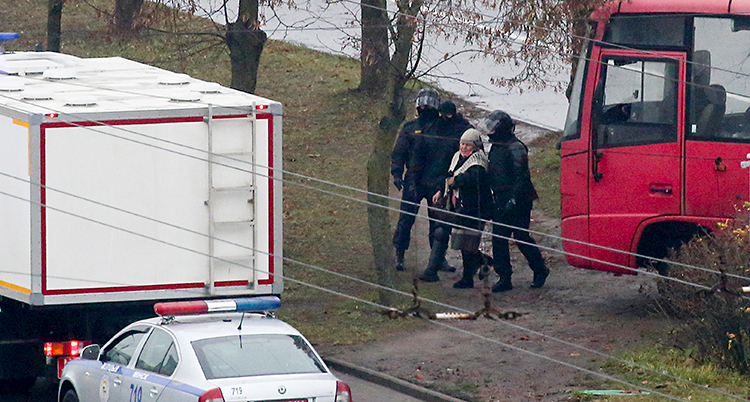 Tre poliser har tagit fast en kvinna. Poliserna har svarta kläder och masker för ansiktet. Runt dem ser vi några träd och några bilar.