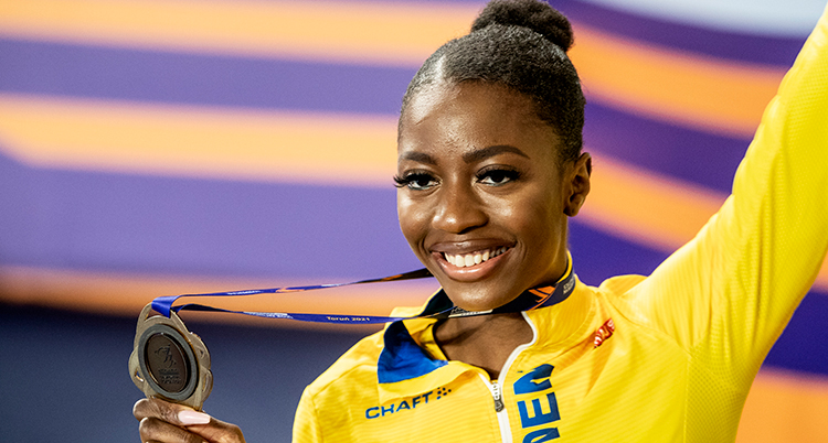 Khaddi Sagnia håller upp medaljen med höger han. Vänster arm sträcker hon i luften. Han har gul jacka. Hon ler och ser glad ut.