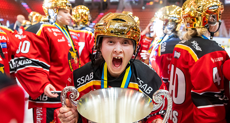En spelare håller fram en pokal och skriker. Hon har en hjälm i guldfärg. Bakom henne är flera spelare med likadana hjälmar.