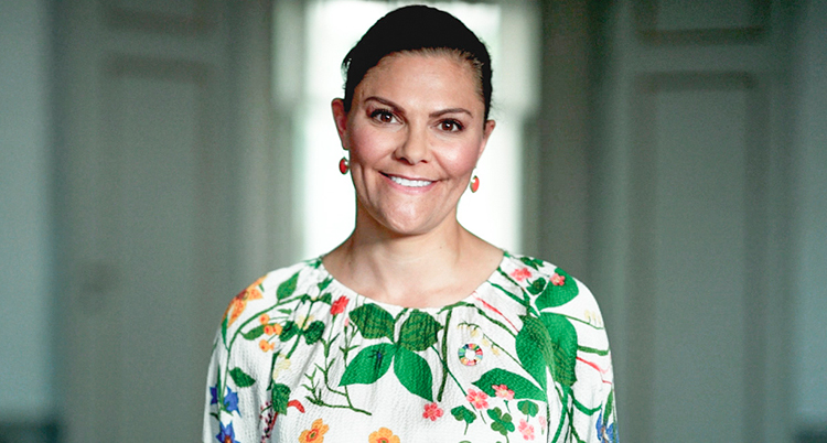 Hon har en blommig blus, mörkt uppsatt hår och tittar rakt in i kameran.