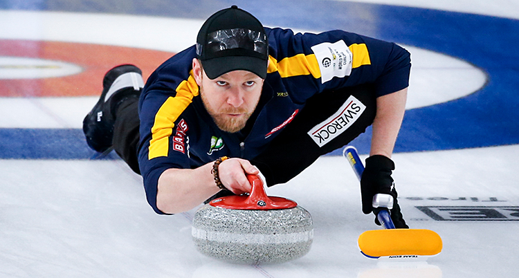 Bilden är från en match i curling. Vi ser Niklas Edin på bilden. Han ligger på isen och siktar med en sten. Han har en keps på huvudet.