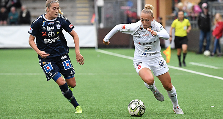 Från en match i fotboll. En spelare i vita kläder springer med bollen. En spelare i blå kläder försöker ta bollen.