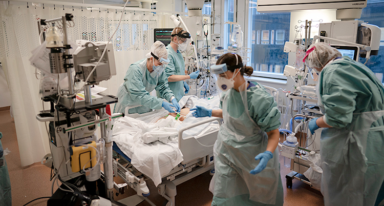 Från ett rum på sjukhus. Fyra personer jobbar. De har skyddskläder och skydd för ansiktet. På sängen ligger en patient. Patienten är skymd, så ansiktet syns inte.