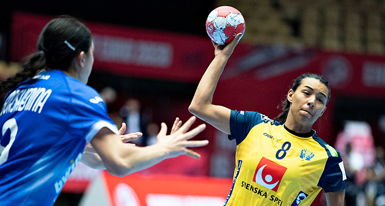 Från en match i handboll. En kvinna i Sveriges lag håller i bollen. Hon hoppar med bollen och ska skjuta ett skott. Hon har en gul tröja. En motståndare står bredvid. Hon har en blå tröja.