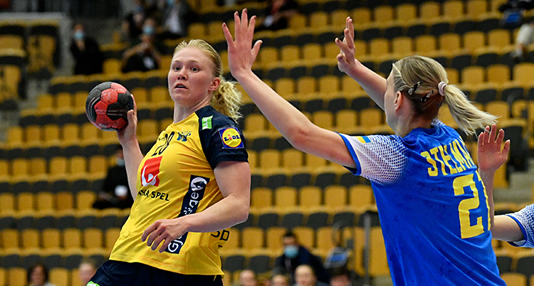 Från en match i handboll. En kvinna med gul kvinna håller bollen i sin högra hand. En kvinna med blå tröja försöker stoppa henne.