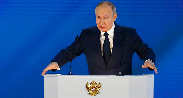 Vladimir Putin står i en talarstol. Han pratar i en mikrofon. Han har kostym och slips på sig. Det är en blå vägg i bakgrunden.