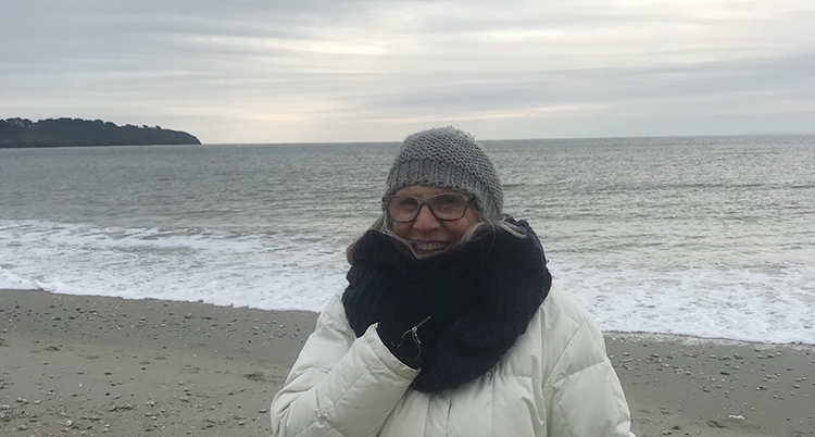 Hon står vid havet. Hon har mössa och halsduk på sig.