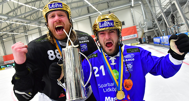 Två spelar höjer varsin näve och vrålar. De har hjälmar av guld och håller i en pokal.