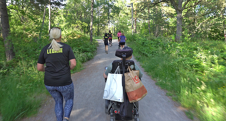 Flera personer går och springer på en väg i en skog. En person åker i en rullstol. Vi ser personerna bakifrån.