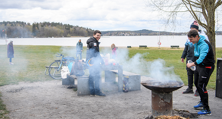 Några personer står vid en eld. Det ryker från elden. En sjö syns i bakgrunden.