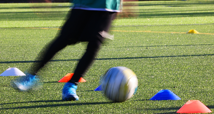 En ung person driver enn fotboll mellan små koner på en fotbollsplan. Bara benen och bollen syns.