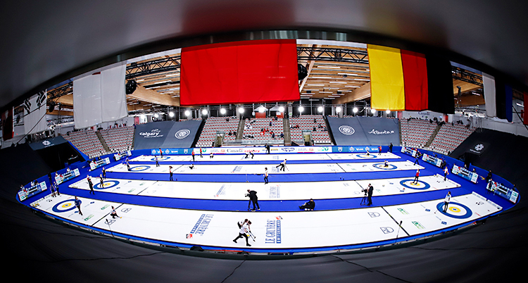 Bilden visar en stor hall för curling. Det är flera banor på is. Lag spelar matcher på de olika banorna.