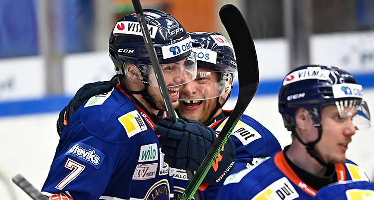 Från en match i ishockey. Tre spelare firar ett mål tillsammans. De är glada. De har hjälmar, klubbor och blå dräkter.