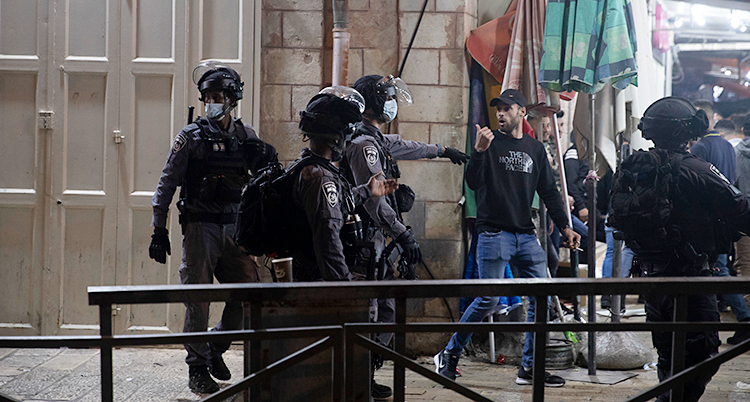 Från en gata i staden Jerusalem. Fyra poliser försöker få bort en ung man från platsen. En av poliserna knuffar på mannen. Poliserna har hjälmar på sig. Mannen har en keps.
