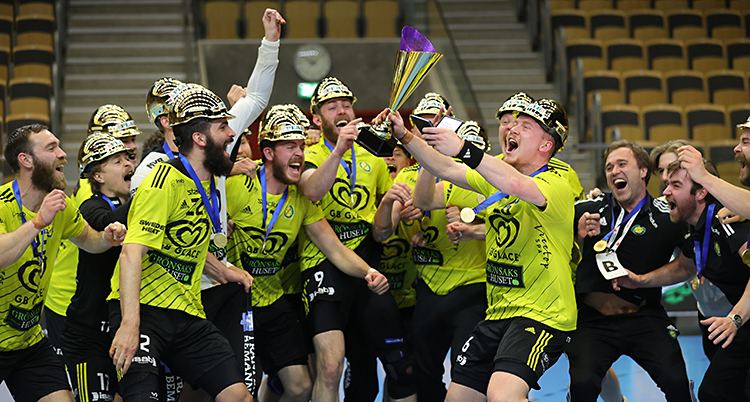 Bilden är tagen i en sporthall. Flera män står tillsammans och jublar. De håller i en pokal. De har gula tröjor och guldhjälmar på huvudet.