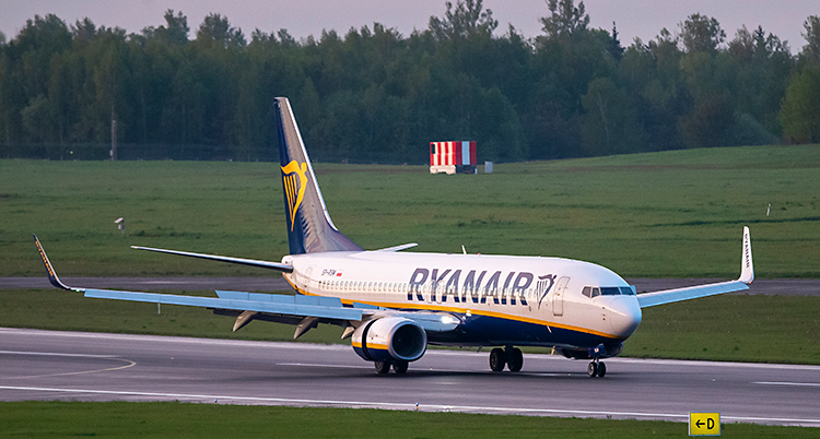 Ett flygplan har precis landat på en flygplats. Det står Ryanair på flygplanet. Bredvid landningsbanan finns ett grönt fält. Längre bort syns en skog.