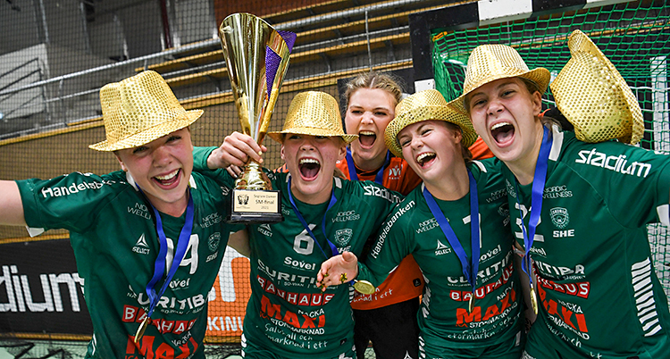 Efter en match i handboll. Fem spelare är glada och jublar. De har guldhattar på sig. En av spelarna håller i en pokal.