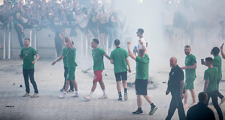 Några fotbollsspelare står utanför en arena. Runt dem finns staket. Bakom staketen finns folk som hyllar dem. Det är rök i luften.