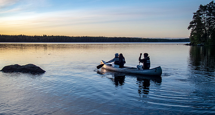 Solnedgång över en stilla sjö. tre personer i en kanot paddlar över sjön.