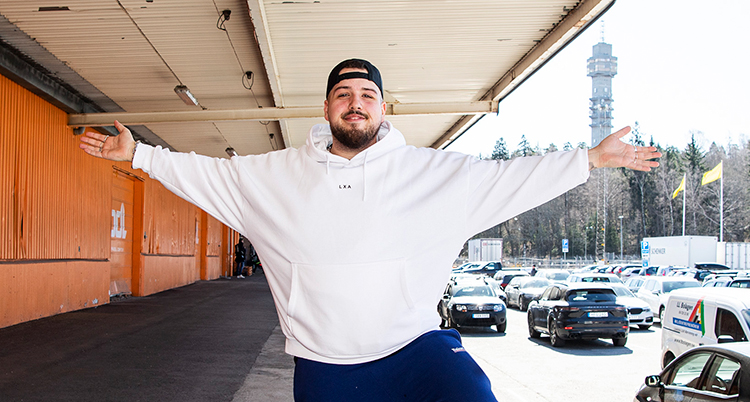 Han har en vit hoodie, bakvänd keps och mörkt skägg. Han sträcker ut armarna och ler in i kameran. Han står utomhus och i bakgrunden syns Kaknästornet.