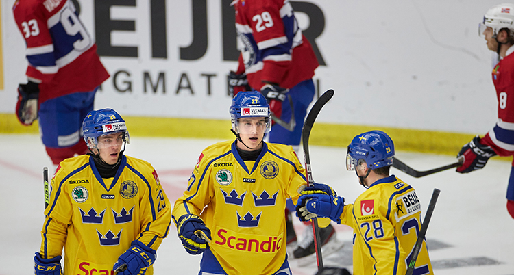 Tre ishockeyspelare i gula landslagströjor står i bredd på en is inomhus. De har blå hjälmar. I bakgrunden syns motståndare i röda tröjor.
