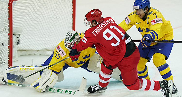 Från en match i ishockey. En spelare i det ryska laget är vid Sveriges mål. Den ryska spelaren försöker göra mål. Den svenska målvakten och en annan svensk spelare försöker stoppa ryssen.
