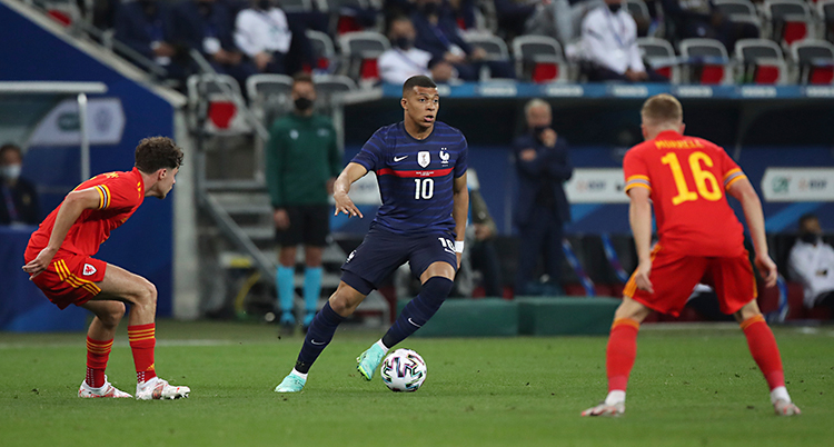 Från en match i fotboll. Kylian Mbappé har bollen och försöker ta sig förbi två motståndare. Han har blå kläder.