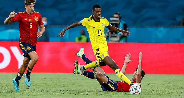 Från en match i fotboll. Alexander Isak har bollen. Han har gula kläder. Två spanska spelare är runt honom. De har röda kläder.