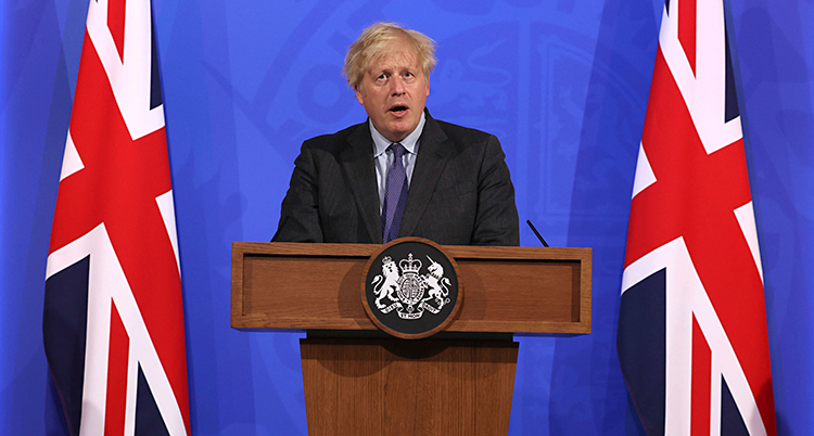 Boris Johnson står och håller ett tal. Han står i en talarstol och pratar i en mikrofon. Han har spretigt ljust hår och en kostym. Bredvid står Storbritanniens flagga.